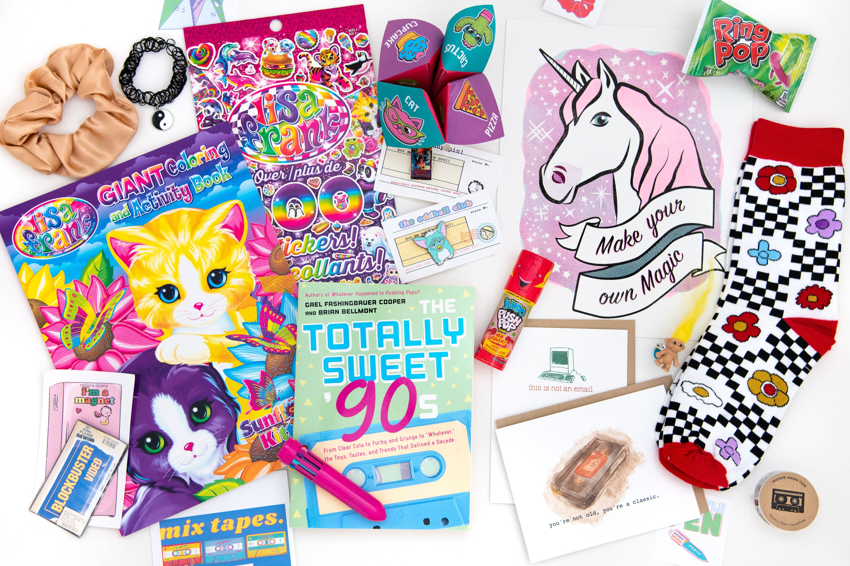 A photo of oddball club 90s box contents including Totally Sweet 90s coffee table book, lisa frank coloring and activity book,  cootie catcher in bright colors, lisa frank stickers, unicorn art print that says make your own magic, two greeting cards that read: 1. this is not an email with a retro computer 2. tou're not old you're classic with a retro watercolor vhs tape. There is also a pair of checker socks, ring pop candy, tape deck washi tape, blockbuster magnet, scrunchi, and yin yang stretch necklace. 